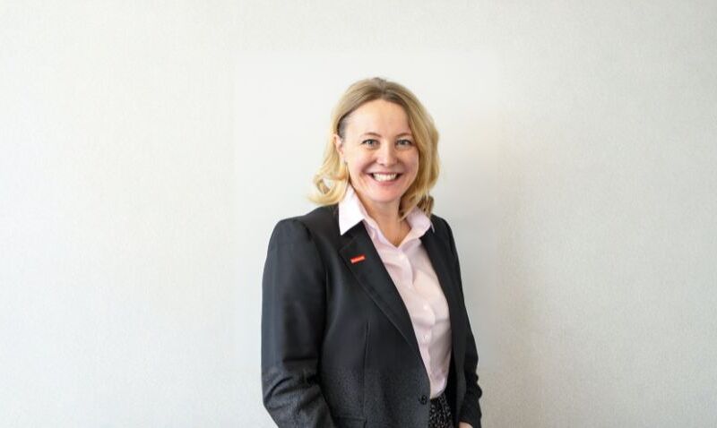 Eine lächelnde Frau in einem schwarzen Blazer mit einem rosa Hemd steht vor einem hellen Hintergrund.